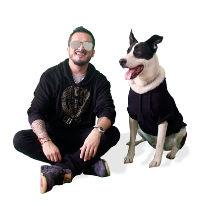 Perro de raza grande negro y blanco con sudadera con gorro negra y diseño dorado y humano con sudadera que combina.